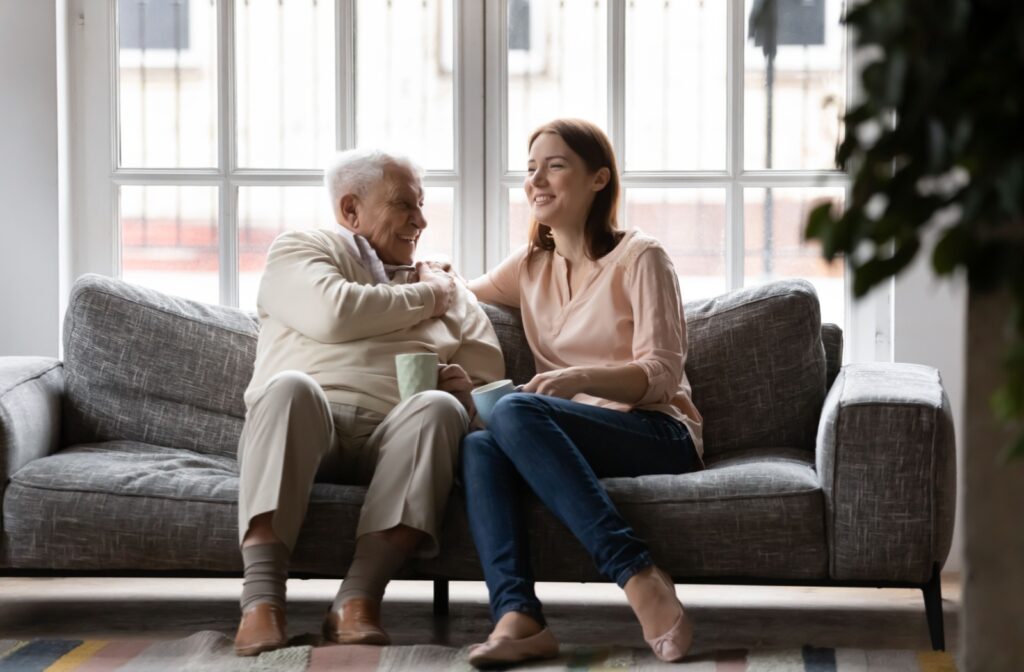 An adult visiting their older parent in senior living, laughing while they chat over coffee on the couch.