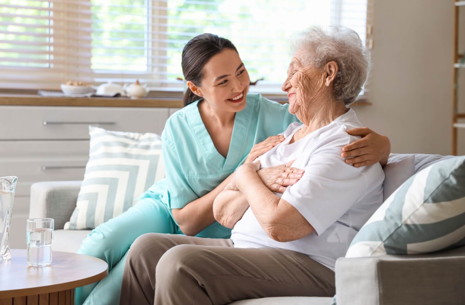 A caregiver in scrubs hugs a happy senior on a sofa as a way of showing support for their healing process in respite care